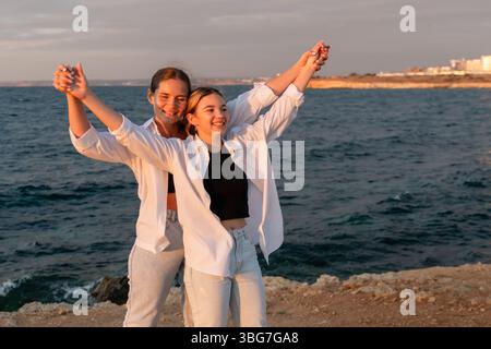 Paare Beach Sunset Happiness: Zwei Menschen in weißen Hemden stehen bei Sonnenuntergang an einem Strand mit erhobenen Armen in der Luft. Stockfoto