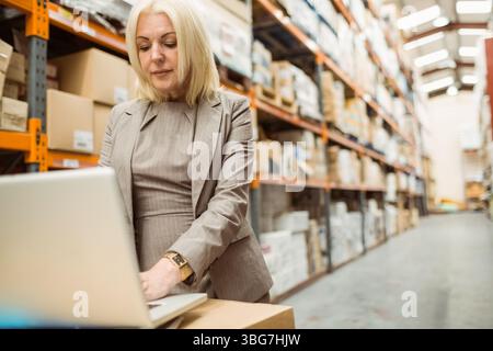 Frau im Anzug überprüft den Bestand im Lagerhaus mit Laptop neben Kisten auf Regalen, Kopierraum Stockfoto