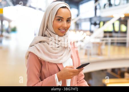 Frau mit beigefarbenem Kopftuch und rosa Blazer, die Smartphone hält und im Atrium des Einkaufszentrums lächelt Stockfoto