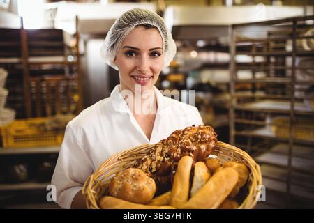 Bäckerin, die in der Bäckerküche neben Metallregalen einen Korb mit frischem Brot hält Stockfoto