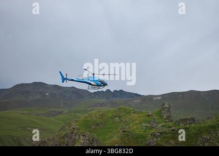 Der Hubschrauber schwingt über einer lebendigen grünen Landschaft, umgeben von felsigen Bergen unter einem grauen Himmel. Die Szene fängt einen Moment der Lufterkundung in A n ein Stockfoto