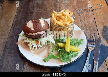 Moving Mountain Vegan Burger mit Brezelbrötchen und veganem pattie, Salat und Sauce, serviert mit einem Korb mit Pommes Frites, Rucoleblattsalat und Dill-Gurken Stockfoto