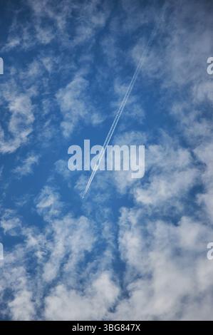 Jet sieht einen weißen Kondensstreifen vor dem Hintergrund eines klaren blauen Himmels voller weicher, flauschiger Wolken. Helles Sonnenlicht verbessert die Farben und A Stockfoto