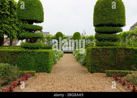Ein formeller Garten mit ordentlich geschnittenen Hecken und Steilbäumen, die einen Kiesweg säumen, der zu einer weißen Bank führt. Stockfoto