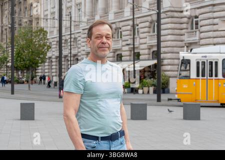 Porträt eines erwachsenen Mannes 50-55 Jahre alt auf einer Stadtstraße. Ein Mann in einem hellen T-Shirt steht vor dem Hintergrund des klassischen europäischen Archits Stockfoto