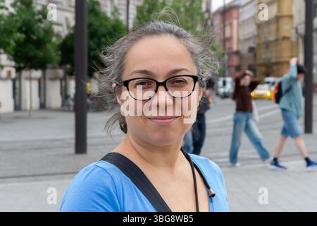 Porträt einer lächelnden 40-45-jährigen Frau, die eine Brille auf einer Straße trägt. Eine Frau mittleren Alters mit natürlichem Aussehen und selbstbewusstem Lächeln posiert Stockfoto