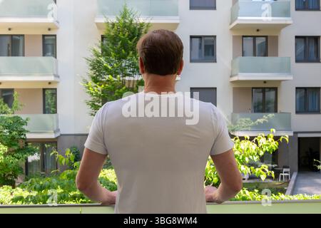Ein Mann in einem weißen T-Shirt steht auf einem Balkon mit Blick auf einen Wohnblock und genießt die Aussicht und einen Moment der Einsamkeit Stockfoto