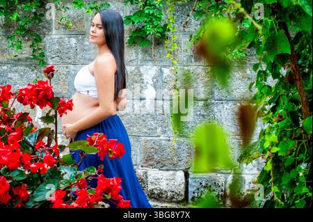 Seitenansicht einer schwangeren Frau, die in einem üppigen Garten neben einer Steinmauer steht. Sie trägt ein weißes Oberteil und einen fließenden blauen Rock, umgeben von leuchtendem Rot Stockfoto