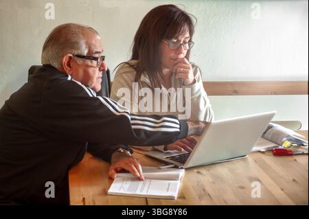 Ein älterer Mann und eine Erwachsene Frau sitzen an einem Holztisch und arbeiten zusammen, während sie sich auf einen Laptop-Bildschirm konzentrieren. Um sie herum sind Dokumente, Stifte, A Stockfoto