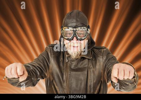 Bildkomposition mit frontaler Oberkörperansicht eines älteren Mannes in brauner, getragener Motorradjacke, Motorradkappe und traditioneller Motorradbrille ho Stockfoto