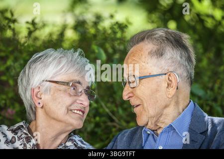 Kopf-Schulter-Porträt eines älteren Rentnerpaares, das sich im Sommer mit einer Brille vor grünen Blättern in die Augen schaut Stockfoto