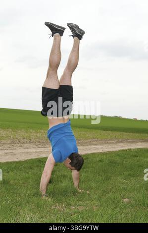 Ganzkörperaufnahme eines jungen Mannes in einem blauen T-Shirt und einer schwarzen Sporthose, der auf einer grünen Wiese und bedecktem Himmel einen Handstand macht Stockfoto