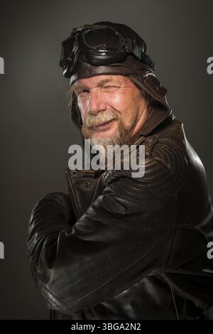 Seitliches Kopf-Schultern-Porträt eines älteren Mannes mit Bart, Lederjacke, Ledermütze und Motorradbrille, die in der Kamera blitzend Stockfoto