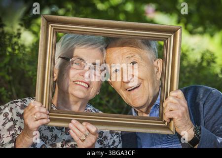 Kopf-und-Schulter-Porträt eines 80-jährigen Rentnerpaares mit Bilderrahmen vor ihren Gesichtern, glücklich posiert vor einem verschwommenen Grün Stockfoto