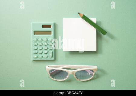 Grüner Taschenrechner und weißer Notizblock, farbiger Bleistift, Brille auf grünem Hintergrund. Draufsicht Stockfoto