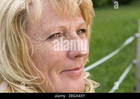 Seitliche Nahaufnahme einer blonden Reifen Frau am Zaun auf einer grünen Wiese, die nachdenklich und zufrieden zur Seite ins Licht schaut Stockfoto