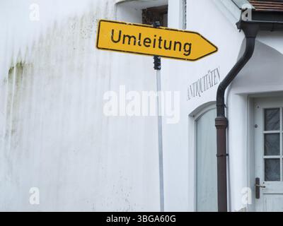 Abgeschrägtes Ablenkungsschild vor einem Antiquitätenladen mit weißer Wand und altem Moos-Putz Stockfoto