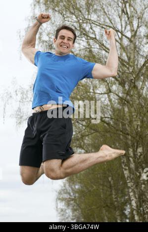 Gefrorene Ganzkörperaufnahme eines jungen Mannes in einem blauen T-Shirt und kurzen schwarzen Sportshorts, die auf einem Sandweg in die Luft springen Stockfoto