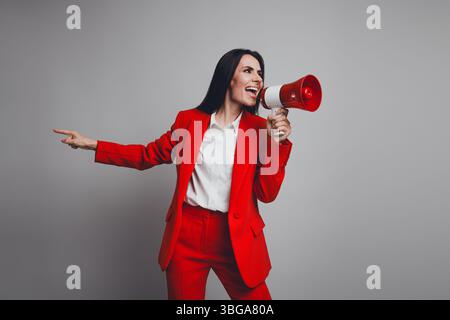 Junge selbstbewusste Geschäftsfrau in trendigem rotem Anzug, die ein Megaphon verwendet, um auf grauem Hintergrund zu kommunizieren und die Szene zu stärken Stockfoto