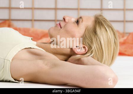 Profiloberkörperansicht einer jungen blonden Frau, die auf dem Boden liegt und die Arme hinter dem Kopf gekreuzt hat Stockfoto