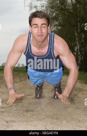 Frontale Ganzkörperaufnahme eines Mannes, der in kurzer blauer Sportbekleidung auf einem sandigen Pfad in einer starken aufwärts gerichteten Liegestützposition zur Kamera zeigt Stockfoto