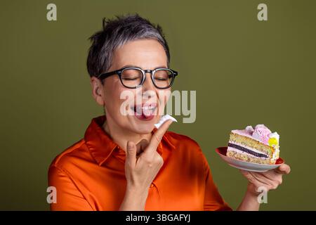 Reife Frau in stilvoller Kleidung genießt ein Stück Kuchen mit Begeisterung und Ausdruck vor einem neutralen Hintergrund Stockfoto