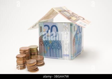 Nahaufnahme eines Hauses aus Euro-Banknoten gefaltet mit davor gestapelten Cent-Münzen auf weißem Hintergrund, Berlin, Deutschland, Europa Stockfoto
