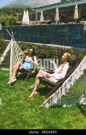 Eine Blondine in weiß und eine Brünette in blauen Kleidern teilen sich tagsüber türkischen Kaffee in einem luxuriösen Hotelgarten. Stockfoto