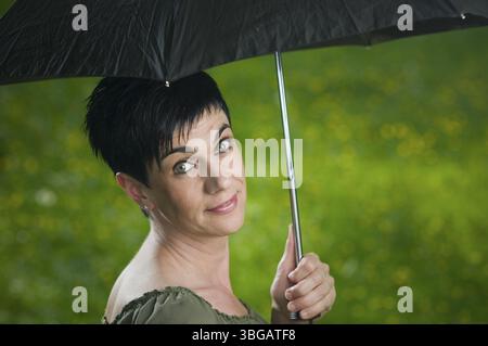 Seitliches Kopf-und-Schultern-Porträt einer kurzhaarigen, schwarzhaarigen Frau mittleren Alters mit einem schwarzen Schirm in einem Park, der freundlich über ihrer Schulter aussieht i Stockfoto
