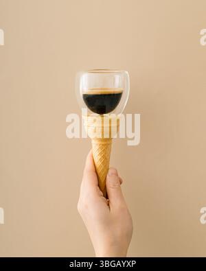 Ein einzigartiges konzeptionelles Foto einer weiblichen Hand, die eine Eiskegel mit einer Tasse Kaffee auf beigem Hintergrund hält. Minimales surreales Essen- und Getränkekonzept. Stockfoto