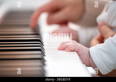 Beschreibung: Nahaufnahme der Hand eines Babys in einem weißen Pullover, der eine Klaviertaste in einer hellen Umgebung berührt. Im Hintergrund spielt eine Erwachsene Hand Klavier. Stockfoto