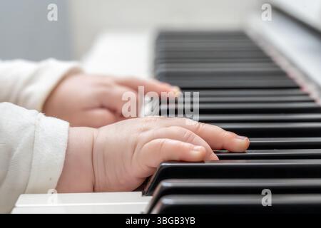 Beschreibung: Super Nahaufnahme der Hand eines Babys in einem weißen Pullover, der das Klavier in einer hellen Umgebung spielt. Junges Leben und Musik. Stockfoto