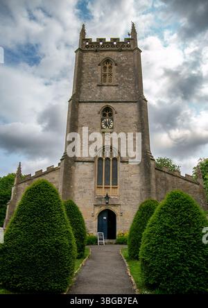 Historische Steinkirche von All Saints Nunney Somerset mit einem hohen Glockenturm und verzierten Fenstern, umgeben von grünen Hecken unter einem dramatischen bewölkten Himmel Stockfoto