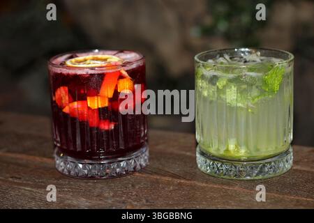 Zwei Cocktails in Gläsern auf einem Holztisch, einer rot mit Obst, der andere Zitrone und Minze. Perfekt für Bars, Sommer oder Erfrischungen. Stockfoto
