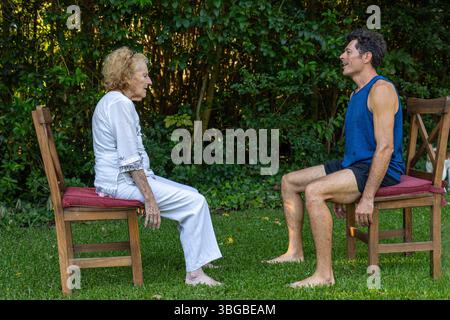 Seniorin und ihr persönlicher Trainer üben Rehabilitationsübungen, sitzen auf Stühlen im Garten, fördern gesundes Altern und einen aktiven Lebensstil Stockfoto