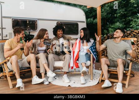 Interrassische Freunde, die Gitarre spielen und singen, amerikanische Flagge halten, Bier trinken, in den Sommerferien in der Nähe von RV sitzen Stockfoto