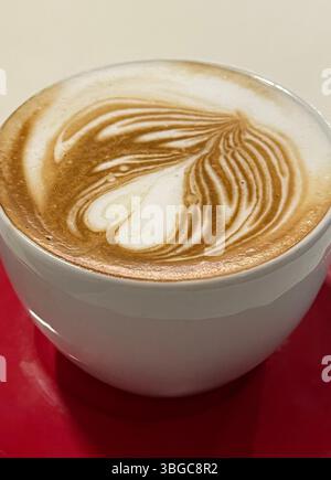 Cappuccino-Tasse mit Herzmuster auf roter Untertasse. Getränke und Essen. Stockfoto