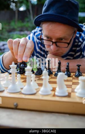 Ein Mann mit gestreiftem Hemd und Hut sitzt tief in Gedanken während eines Freiluftschachspiels. Geistige Herausforderung unter der warmen Sommersonne. Stockfoto