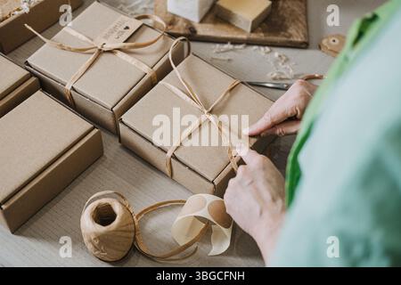 Person bindet Band um Kraftgeschenkboxen auf einem Tisch mit handgefertigten Seifen und Verpackungsmaterialien. Kleinserienproduktion, handwerkliche Wirtschaft, Hersteller Stockfoto