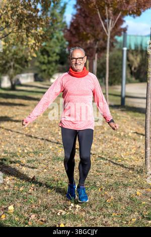 Ein älterer Kaukasier macht im Herbst Dehnungsübungen in einem Park, trägt Brille, ein pinkfarbenes Hemd, schwarze Leggings und blaue Sneakers. Stockfoto