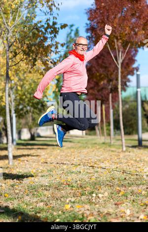 Senior-Kaukasier mit grauen Haaren und Brille, rosa Oberteil, schwarze Leggings und blaue Turnschuhe, springt im Herbst im Park und lächelt Stockfoto