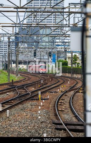 Der rote Pendlerzug fährt auf kurvenreichen Gleisen in städtischer Umgebung. Umgeben von hohen Gebäuden sind die Gleise mit Freileitungen gesäumt. Stockfoto