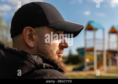 Nahaufnahme eines bärtigen Mannes in einer schwarzen Kappe, mit einem sonnigen Spielplatz, der im Hintergrund verschwommen ist, was auf Freizeit oder Familienzeit hindeutet. Stockfoto