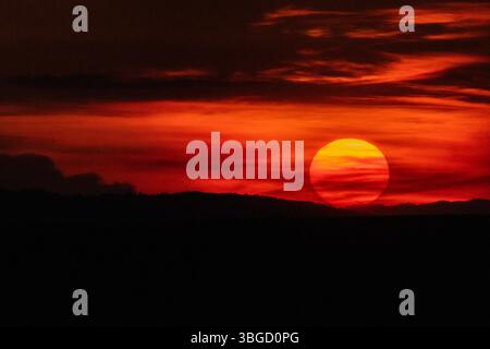 Die Sonne berührt den Horizont und wirft tiefe Karmesintöne über mehrschichtige Wolken und silhouettenreiche Hügel in einer atemberaubenden ländlichen Landschaft bei Sonnenuntergang Stockfoto