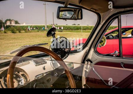 Innenansicht eines Oldtimers mit Holzlenkrad, Armaturenbrettdetails und einem kleinen tragbaren Ventilator, der an einem sonnigen Tag in der Nähe eines Feldes parkt Stockfoto