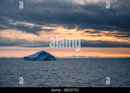 Ein großer Eisberg schwingt unter bewölktem Himmel in der Davis-Straße, mit weit entfernten schneebedeckten Grönlandspitzen, die von der weichen arktischen Dämmerung beleuchtet werden. Stockfoto