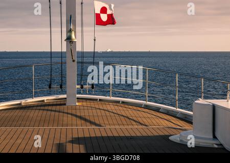 Der Bug eines Schiffes, das die grönländische Flagge auf See vor Disko Banke in Grönland führt, mit ruhiger See und fernen Eisbergen am Horizont. Stockfoto