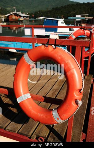Leuchtend orangefarbener Rettungsring an rotem Geländer auf hölzernem Dock mit verschwommenen Booten und Häusern im Hintergrund. Zapadna Morava See in Serbien Land. Stockfoto