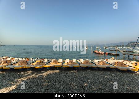 Gardasee, Italien 3. Januar 2025: Tretboote am Kiesstrand des gardasees, nahe bardolino und lazise, mit Segelbooten im Hintergrund; Stockfoto