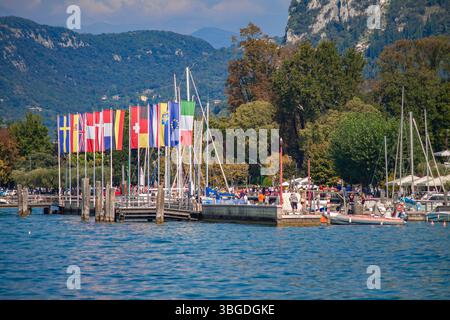 Gardasee, Italien 3. Januar 2025: Europäische Fahnen wehen auf einem Pier mit Segelbooten und Touristen, die den gardasee besuchen Stockfoto